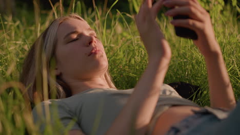 primer plano de una mujer joven acostada en un campo cubierto de hierba, usando el teléfono mientras la luz del sol ilumina suavemente su cara y sus alrededores, con una postura relajada y una mirada enfocada