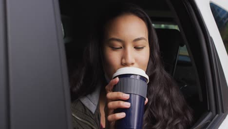 asian woman with lowered face mask looking out of the window and drinking coffee sitting in the car