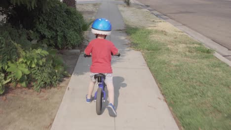 follow shot of a young boy riding a blue balance bicycle along a sidewalk in a residential neighborhood with green yards