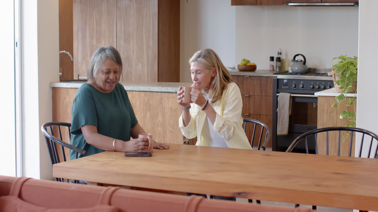 Enjoying coffee, two multiracial senior female friends at table and chatting in kitchen, at home