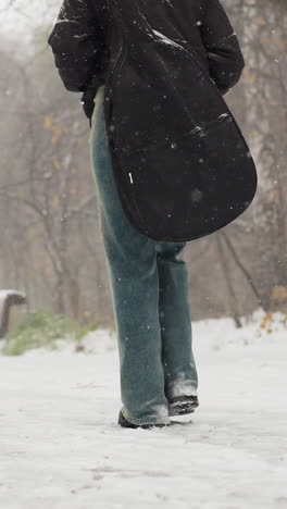 persona que lleva guitarra camina por un camino nevado, otra figura borrosa en la distancia, en medio de árboles de invierno y nieve que cae, capturando una atmósfera serena de invierno