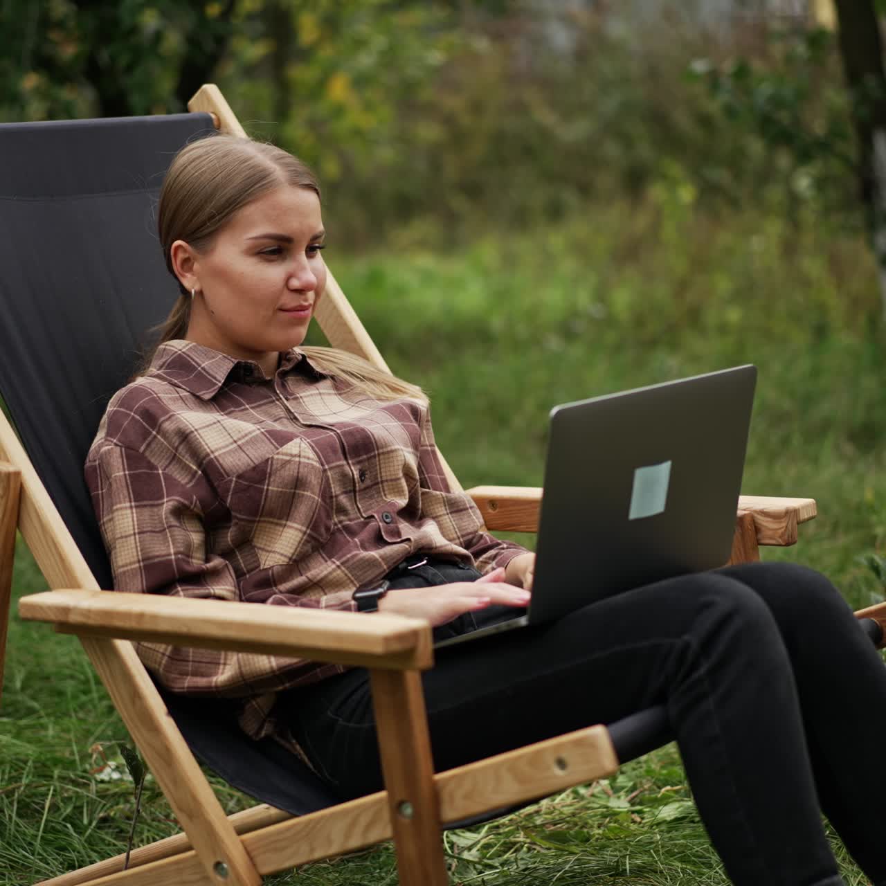 Beautiful woman in chequered shirt starts work on her computer sitting in garden chair. Positive relaxed lady opens laptop and begins typing. Garden at backdrop