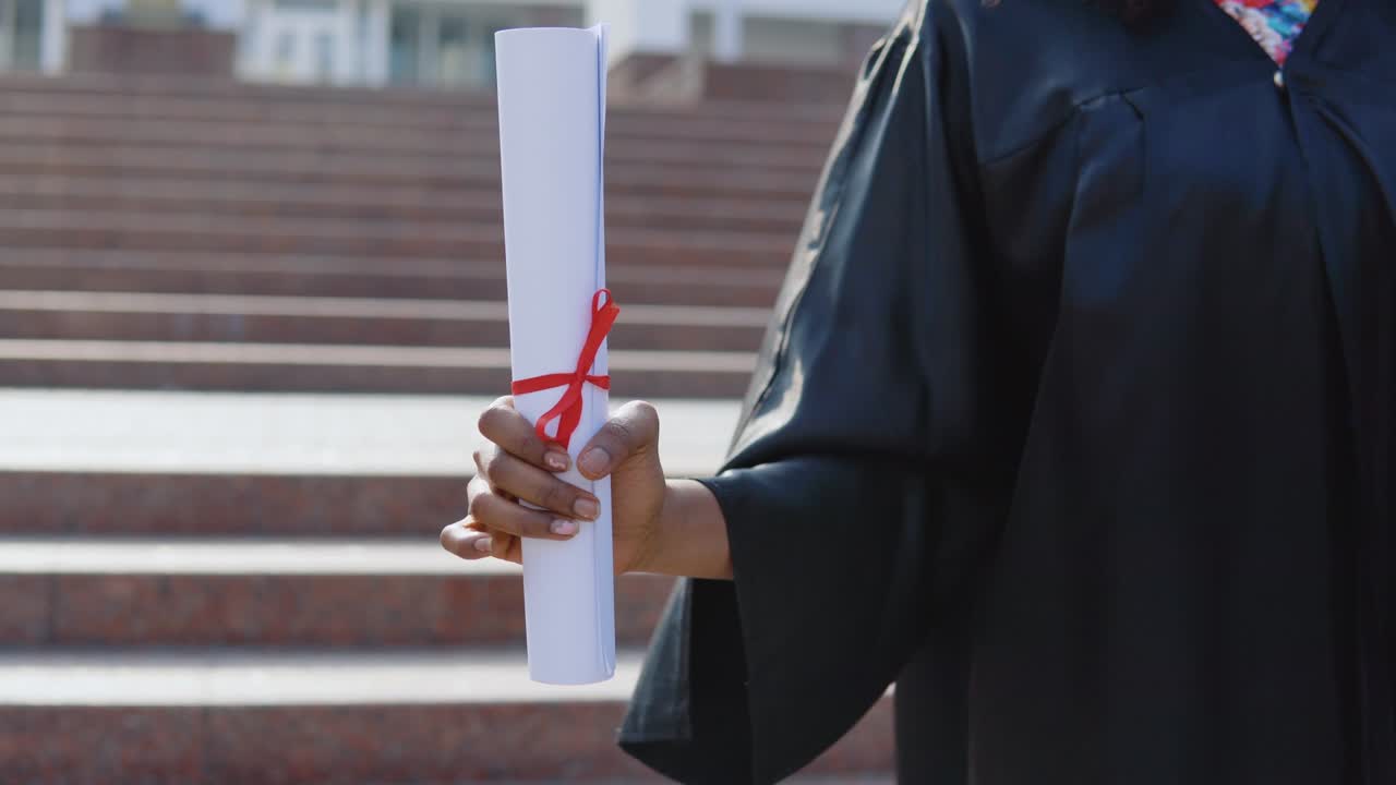 diploma de educación superior verticalmente en las manos de una mujer afroamericana graduada de la universidad en el fondo de las escaleras desde el exterior.