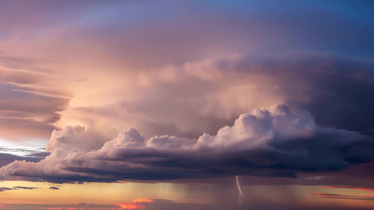 Nubes cumulonimbus masivas se desplazan sobre un atardecer distante, revelando capas esculpidas de luces pálidas y sombras profundas, mientras un estrecho relámpago aparece debajo de la tormenta