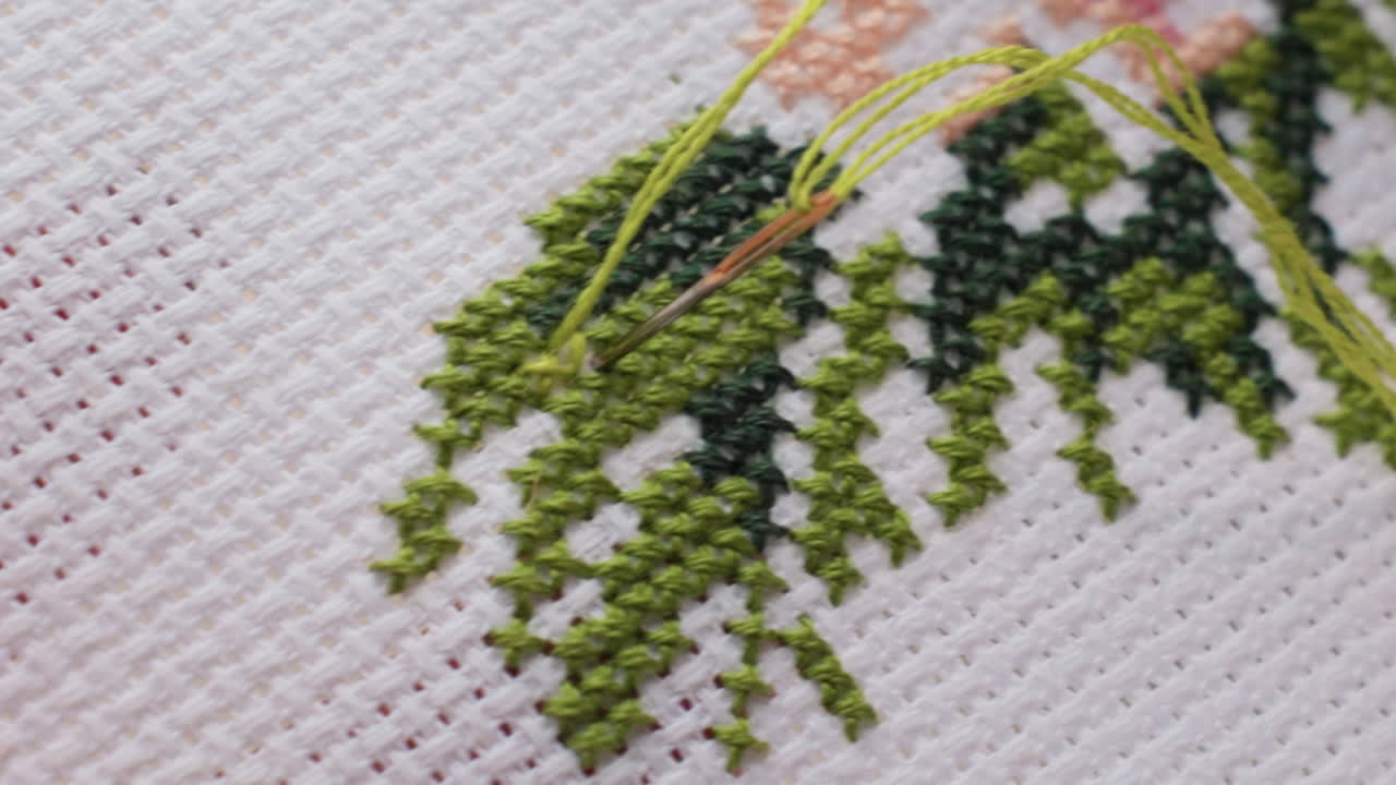 Extreme close up embroiderer guiding green thread through needle into cross stitch leaves on white canvas, blue nail visible, precise hand movement, fine weave texture, calm outdoor daylight mood