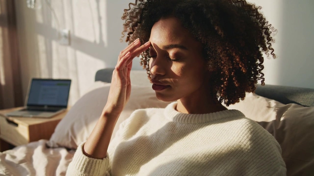 Pausing laptop work, woman wearing cream sweater rubbing temple on bed at home, open laptop visible