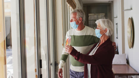 Senior-caucasian-couple-wearing-face-masks-holding-rainbow-painting-against-the-window-at-home