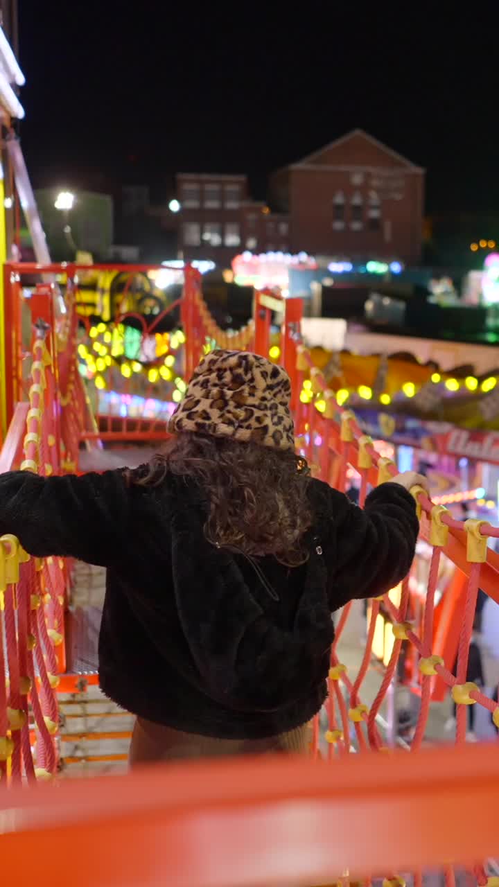 A person with a leopard print bucket hat and furry coat looking out over a brightly lit fairground at night