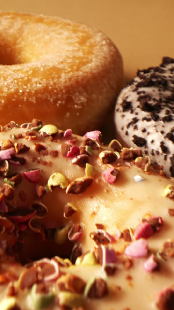 Kinematische Kamerafahrt über verschiedene Gourmet-Donuts mit glänzender Schokoladenglasur, Keksstreusel-Topping und Vanilleguss mit bunten Nussstreuseln. Reichhaltige Texturen und Premium-Backwaren-Optik.