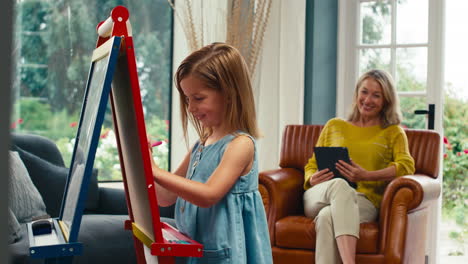 family with daughter drawing picture on easel as mother with digital tablet watches from sofa