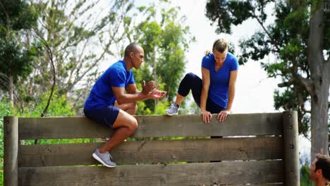 Male-trainer-assisting-fit-woman-to-climb-over-wooden-wall-during-obstacle-course-4k