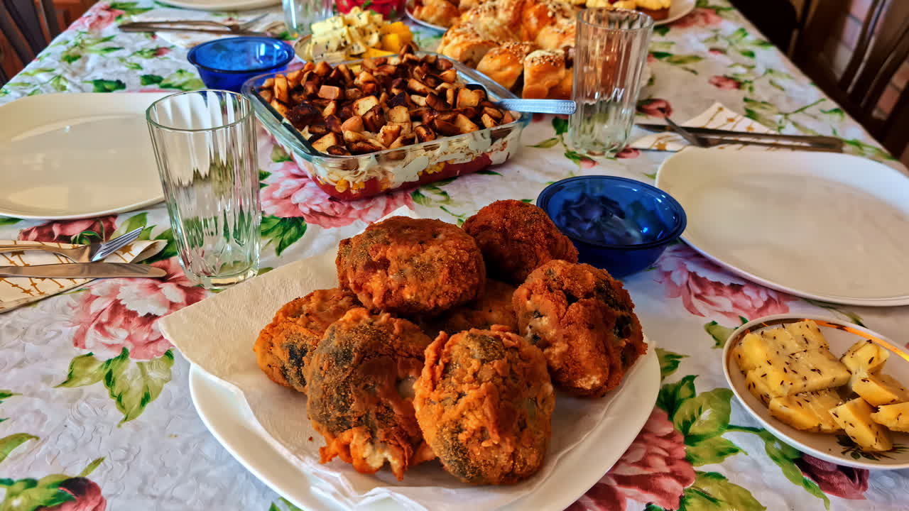 A dolly camera shot reveals a festive table laden with traditional homemade Latvian food, including savory pastries, fried cutlets, roasted potatoes, caraway cheese, and cakes for a celebration