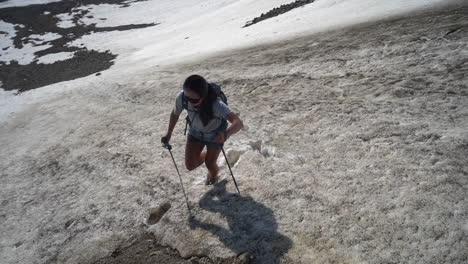 woman with trekking poles walking uphill on snow in mountain landscape on sunny summer day, slow motion