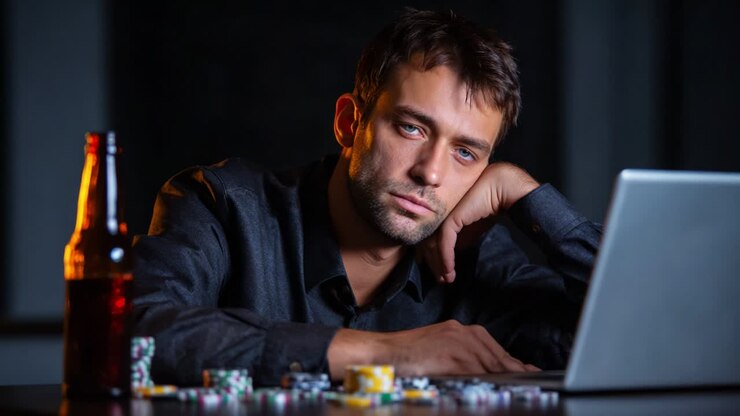 A contemplative man sits at a dimly lit table, gazing intently at his laptop while surrounded by scattered poker chips and a beer bottle, embodying the tension and allure of a high-stakes game