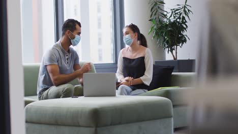 Diverse-male-and-female-business-colleagues-wearing-masks-sitting-on-sofa-making-elbow-bump