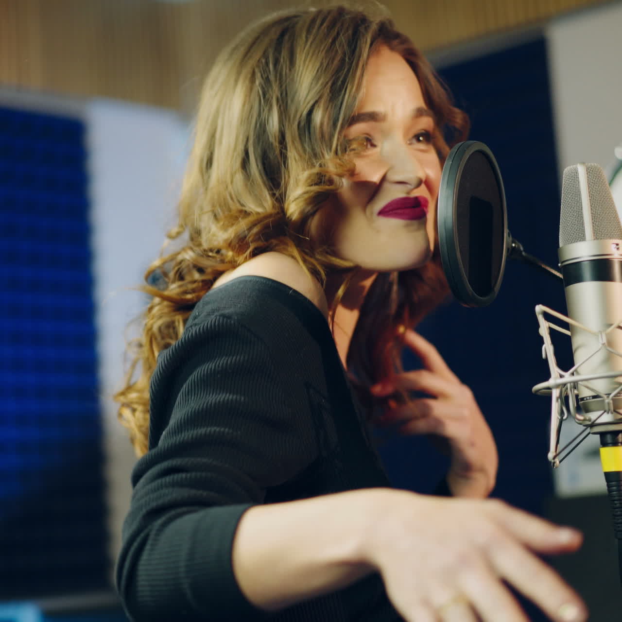 Portrait of a happy female wearing black singing into a modern microphone in audio studio. Side view of a pretty singer and musical equipment