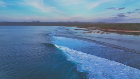 Drone-clip-of-ocean-waves-and-coral-reef-on-shoreline-of-remote-tropical-island-with-thick-jungle-and-pastel-colour-skies