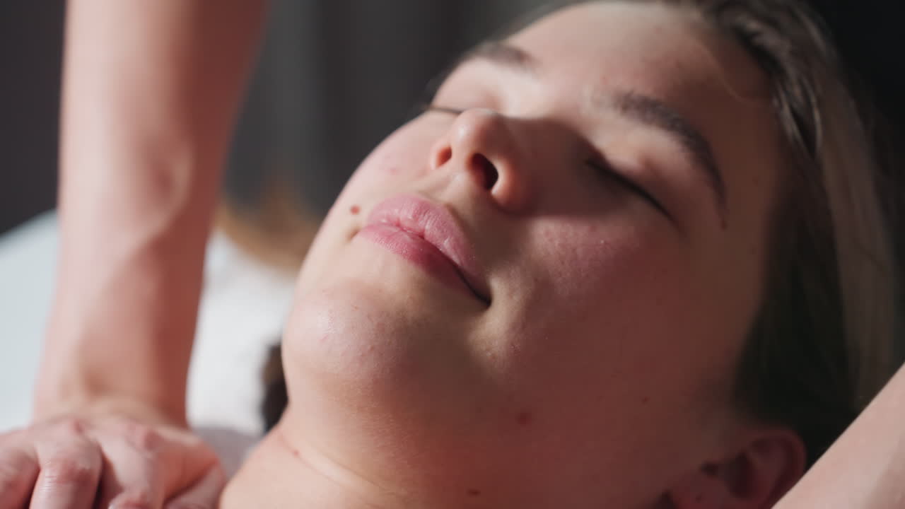 Close up view of light-skinned female client lying with closed eyes and soft smile during massage therapy session, showing relaxed demeanor under gentle touch of therapist s hands