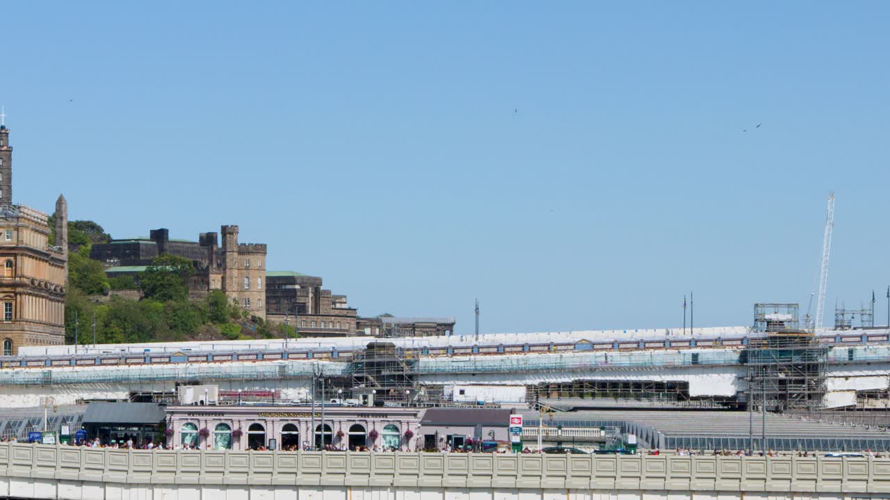 Yellow train enters busy city railway station under clear daylight, wide static shot, urban setting
