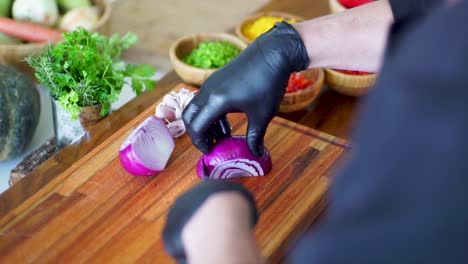 Onions-are-expertly-sliced-on-a-wooden-pad,-hands-protected-by-black-gloves,-while-vibrant-sliced-vegetables-await-in-a-rustic-wooden-bowl