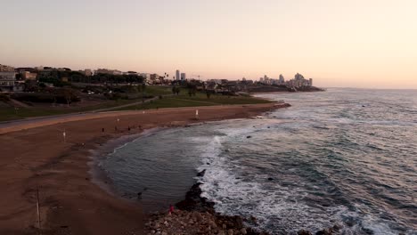 aerial slide to the left drone shot of an empty beach, small waves crashing on the sand, view of a city and a park