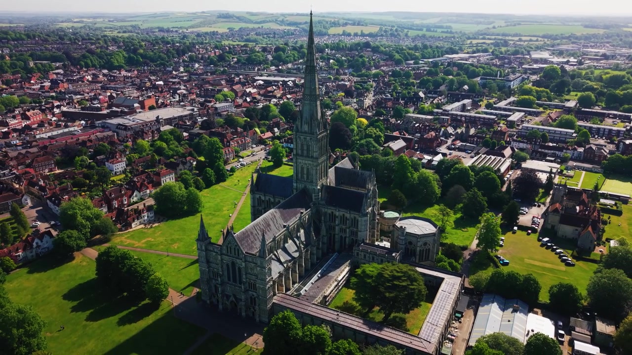 Luftaufnahme mit Drohne über der Kathedrale von Salisbury in Wiltshire, England, die die mittelalterliche gotische Kathedrale, den berühmten Turm, die Rasenflächen des Kathedralenbezirks, Bäume, historische Gebäude und die umliegenden Stadtstraßen zeigt