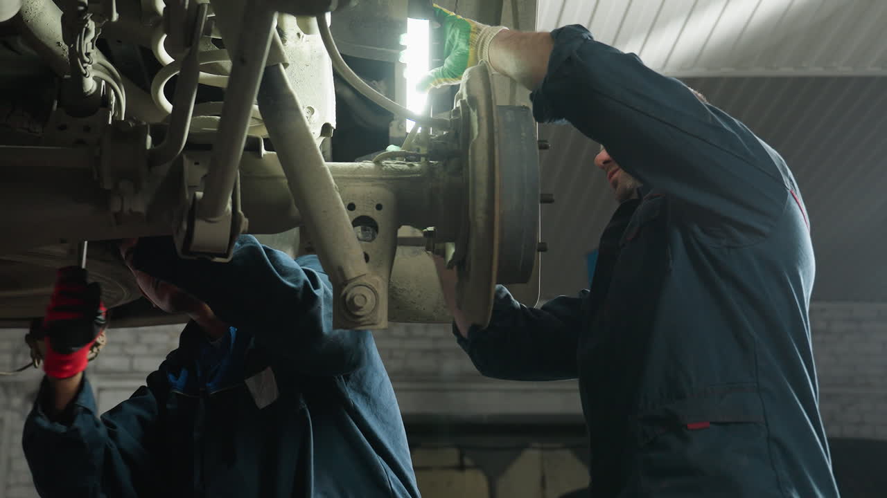 Indian engineer working under car, tightening bolt with focused expression, light reflecting on his face, wearing gloves and blue uniform, engaged in automotive repair inside professional garage
