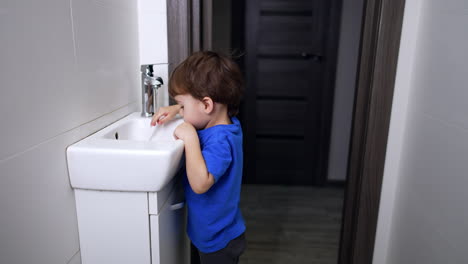 caucasian toddler standing in the bathroom near the sink. curious kid watching the water flow and puts his hand under water.