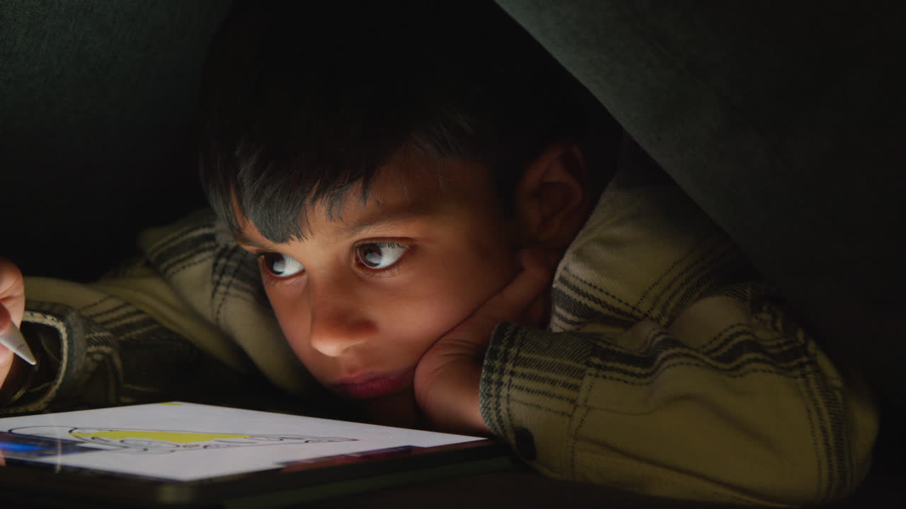 Close Up Of Young Boy In Home Made Camp Made From Cushions Playing And Drawing With Digital Tablet And Stylus At Night 1