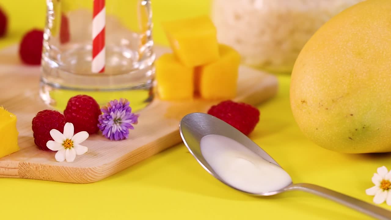 Un cucchiaio pieno di yogurt poggia vicino a un mango intero, lamponi e guarnizioni floreali.