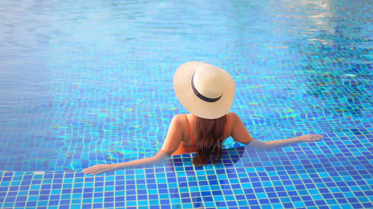 Woman in orange swimsuit and straw cap relaxing in clear warm water of luxury resort swimming pool