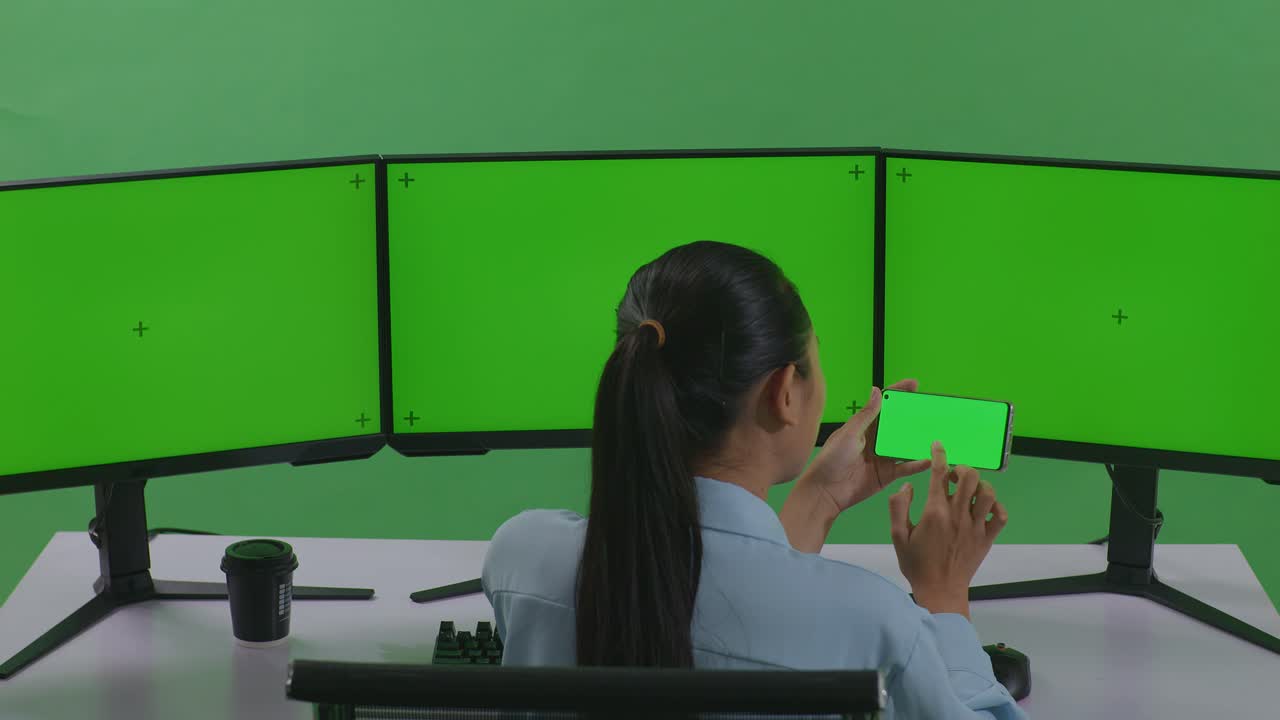 여러 모니터와 함께 녹색 화면 배경에 스마트폰의 사진을 찍는 여성