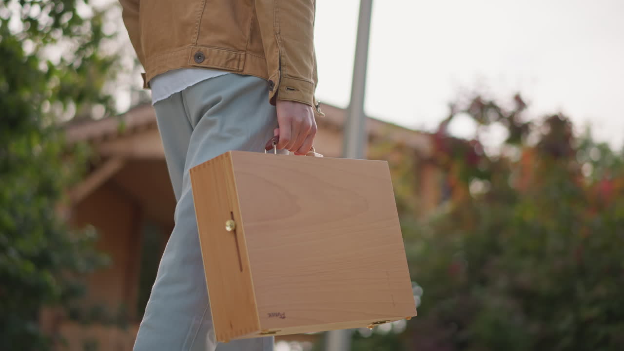 person walking with wooden art case in park, carrying portable sketch box toward suburban porch, jeans and jacket visible, warm sunlight and greenery create calm focused mood for outdoor painting