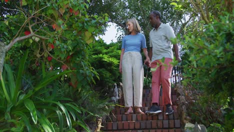 una pareja mayor diversa caminando en su jardín al sol tomándose de las manos y hablando