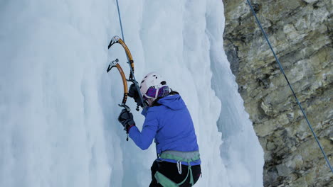 female ice climber hits ice axes into vertical ice cliff as she ascends