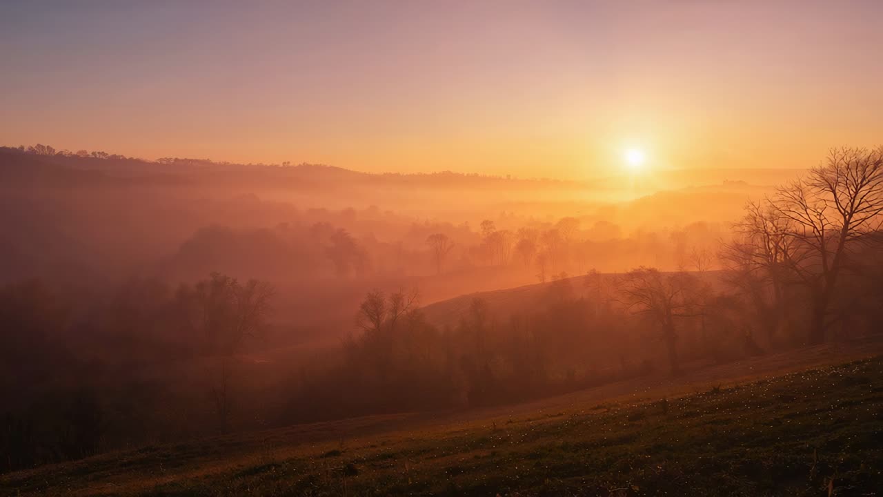 Opkomende zon en optrekkende mist onthullen gelaagde heuvels boven landelijke vallei bij zonsopgang, met grazige helling.