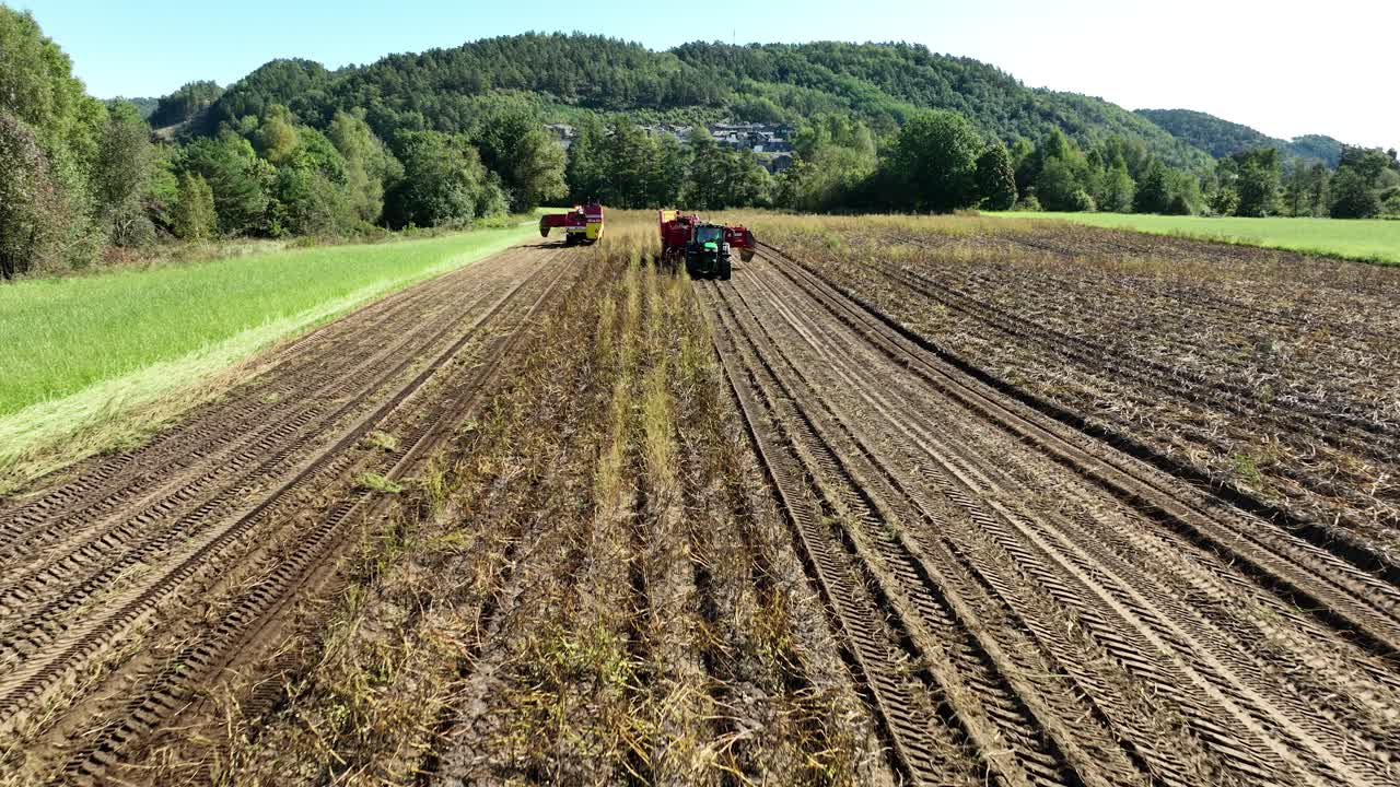 Two large tractors work together in a field, harvesting potato and leaving distinct tire tracks in the soil. Lush green trees and hills provide a backdrop under clear skies