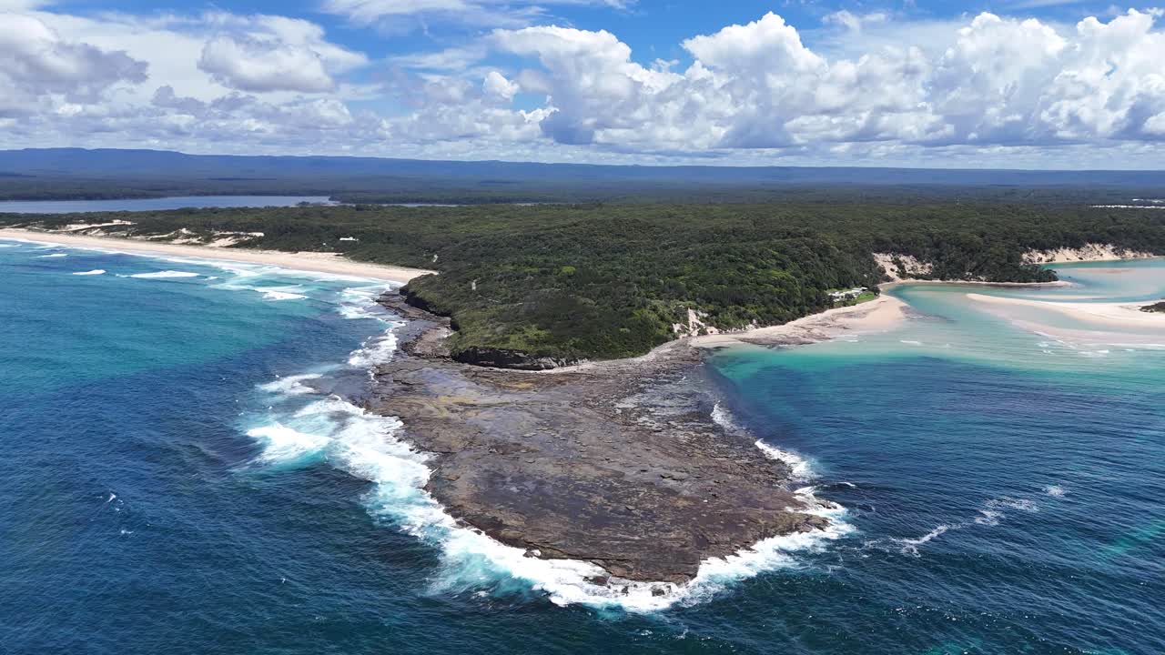 Drone approach over rocky peninsula of Sussex Inlet, NSW, Australia, with deep blue water meeting golden sandy shorelines