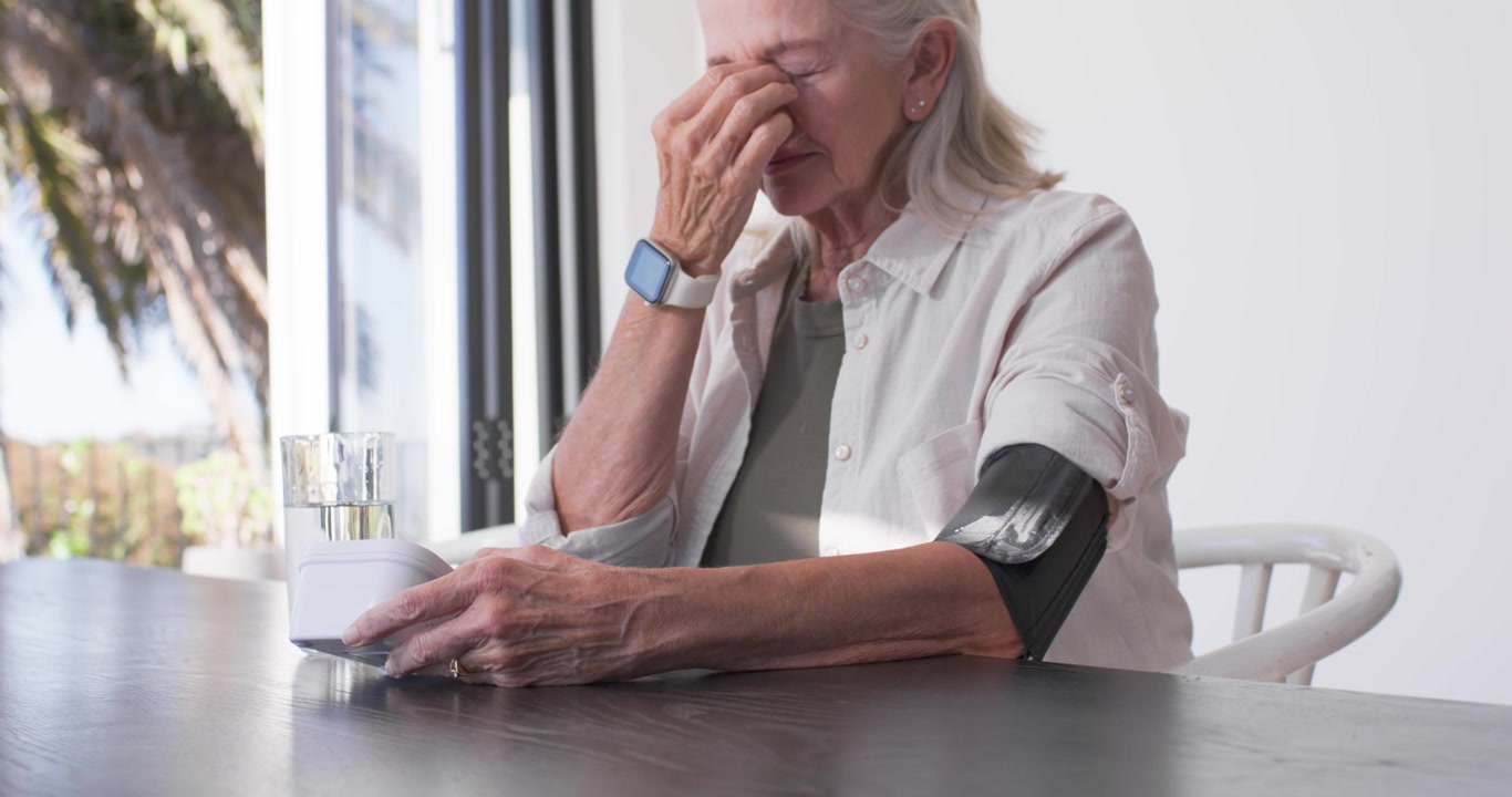 Mujer mayor sentada en una mesa junto a una puerta de cristal, apoyándose en el monitor, frotándose la nariz y revisando el manguito del brazo.