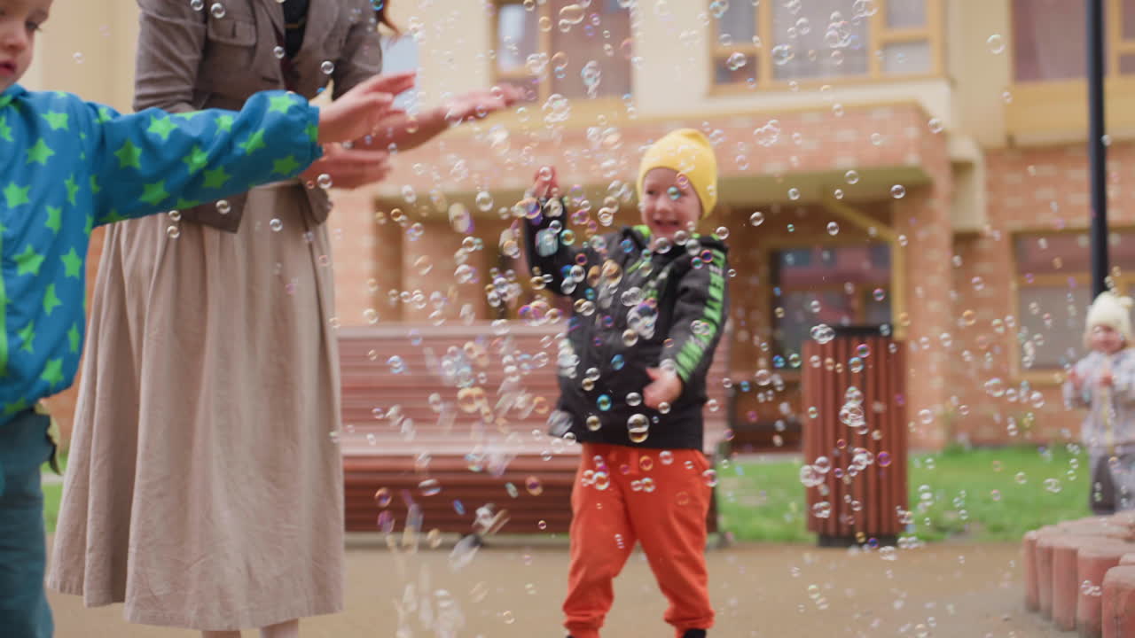 Smiling mother and children standing outdoors in courtyard watching colorful bubbles float from soap machine, showing family excitement, playful curiosity, and joyful bonding