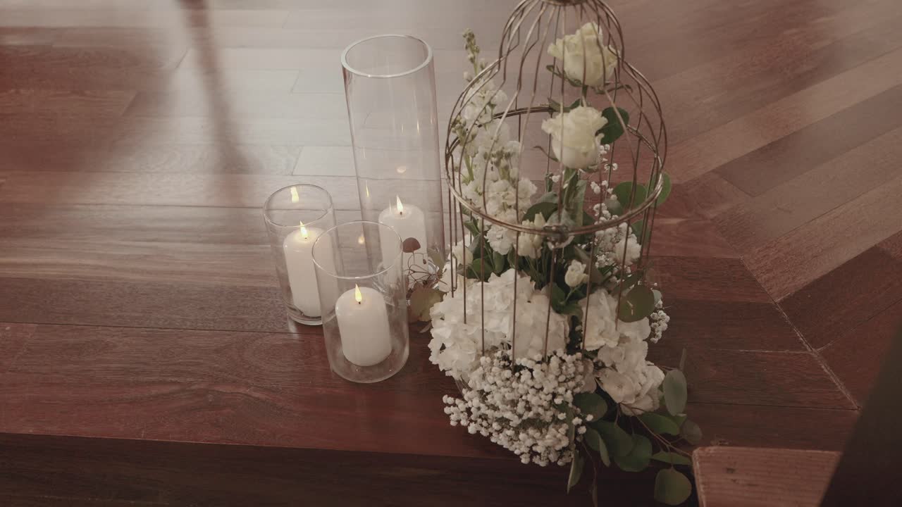 white roses and baby’s breath in birdcage beside glowing candles on dark wooden floor