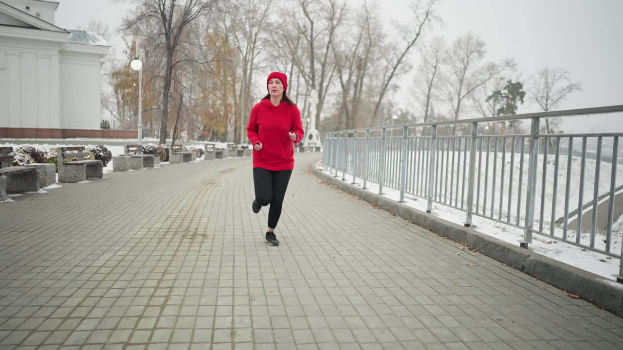 Donna di mezza età che fa jogging lungo un sentiero innevato del parco, indossando una felpa rossa con cappuccio e leggings, concentrata sull'allenamento, circondata da un sereno paesaggio invernale, panchine, ringhiera in ferro, alberi e una borsa lontana su una panchina