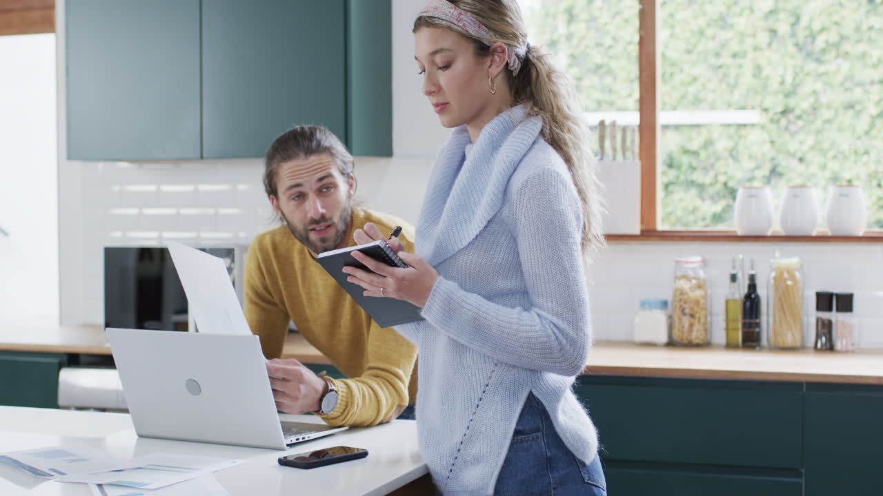 Diverse couple using laptop and taking notes in kitchen at home, in slow motion