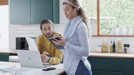 pareja diversa usando computadora portátil y tomando notas en la cocina en casa, en cámara lenta