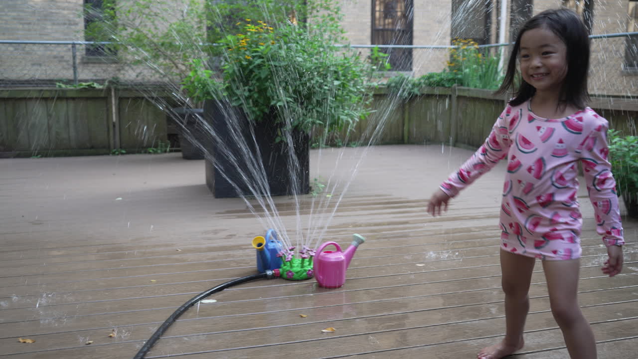 View of a little girl watching a water sprinkler start in the garden on the rooftop with blue and a pink watering can around it