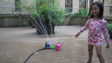 Vista-De-Una-Niña-Viendo-Cómo-Comienza-Un-Rociador-De-Agua-En-El-Jardín-En-La-Azotea-Con-Una-Lata-De-Agua-Azul-Y-Rosa-A-Su-Alrededor