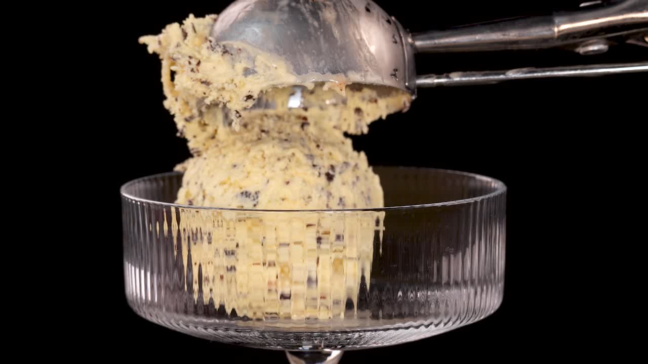 A metal scoop releases a ball of vanilla ice cream with chocolate chips and brownie pieces into a clear glass dessert bowl against a black background, under bright studio lighting