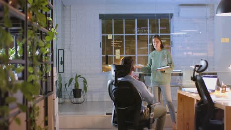 asian casual businesswoman using tablet presenting to male colleague at night in office