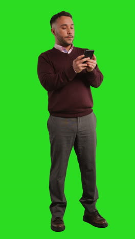 vertical video full length studio shot of businessman using mobile phone to message or browse online standing against green screen