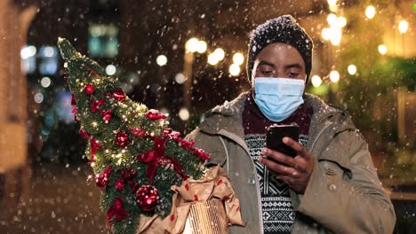 retrato de un joven afroamericano con máscara facial escribiendo en un teléfono inteligente y sosteniendo un árbol de navidad en la calle mientras nieva en navidad